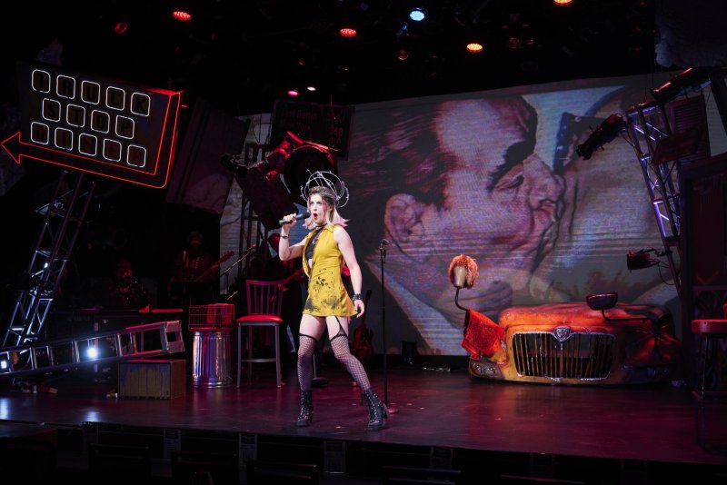 Performer in yellow dress, fishnets, and elaborate headpiece sings on stage with a band, neon sign, and projected image.