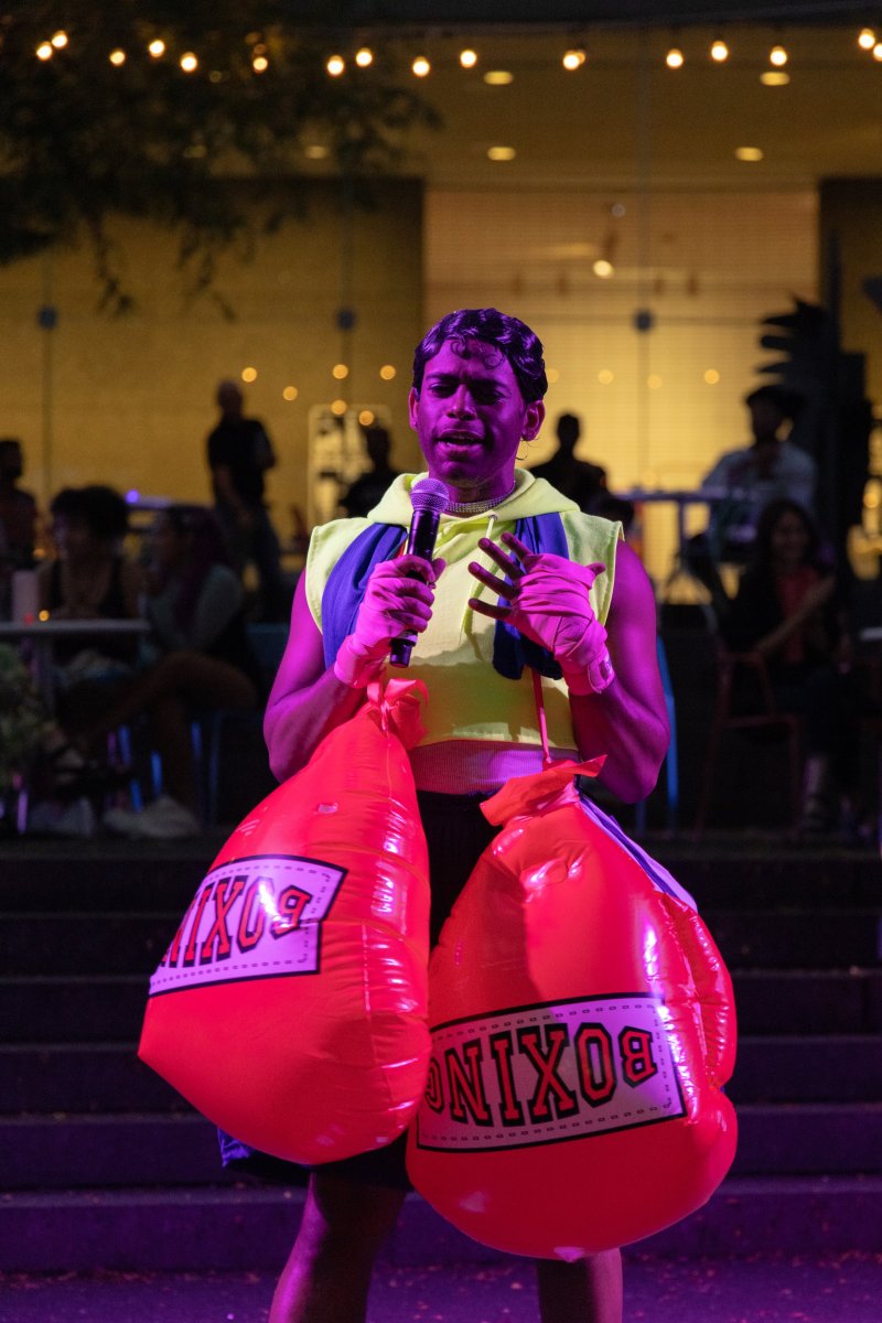 Performer on stage with a microphone, wearing a yellow hoodie vest, pink gloves, and carrying two large inflatable red boxing gloves. Audience is blurred in background.