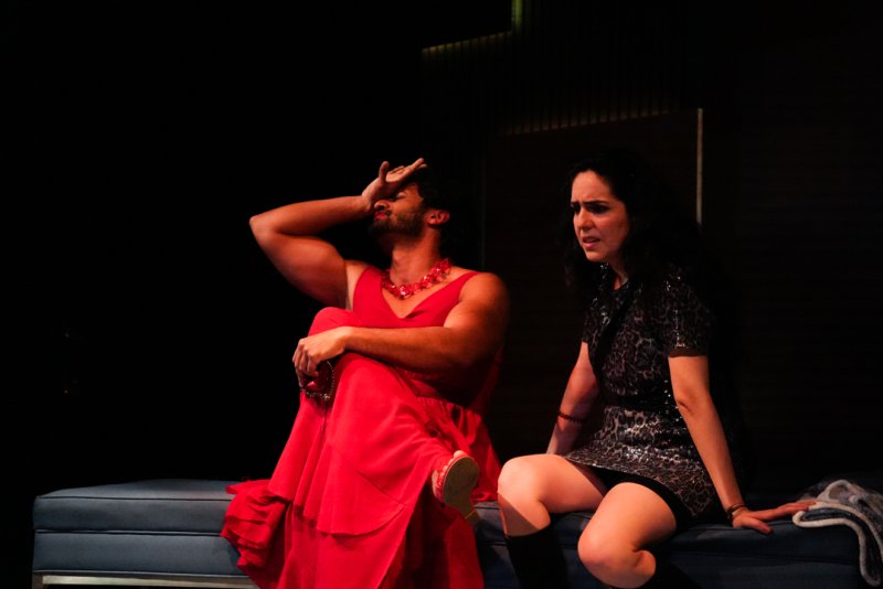 Two people on a bench against a dark background: one in a red dress with hand to forehead, the other in a sparkly dress looking distressed.