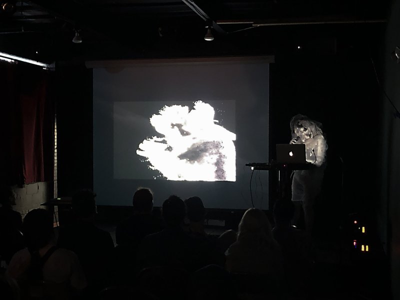 Dark room with audience watching a video projection of a bright white, abstract form. A figure stands to the right, illuminated by a laptop screen.