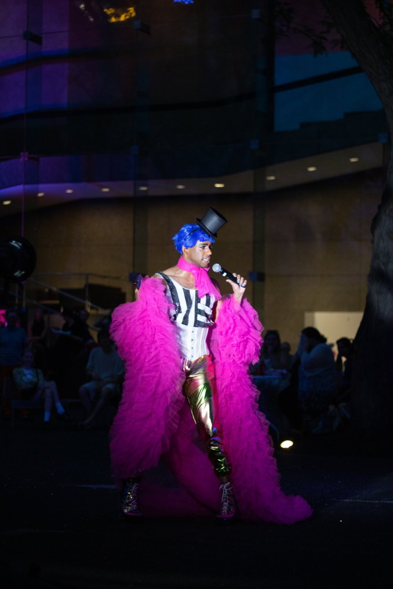 Performer with blue hair and top hat sings into a microphone, wearing a pink ruffled robe, striped top, corset, and gold pants. Audience visible in the background. Stage lighting is blue and purple.
