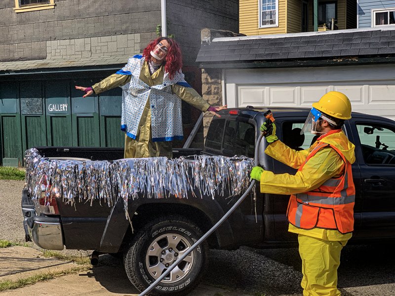 Outdoor scene: person in yellow safety gear sprays water at a clown-like figure standing in a pickup truck bed decorated with silver fringe. Buildings in background.