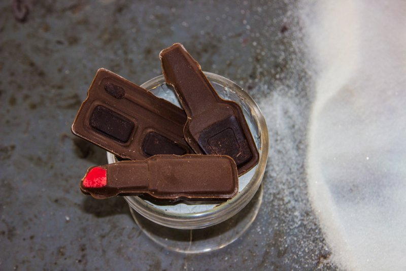 Overhead shot of three brown chocolates shaped like a cassette tape, a bottle, and a lipstick with red tip, in a glass.