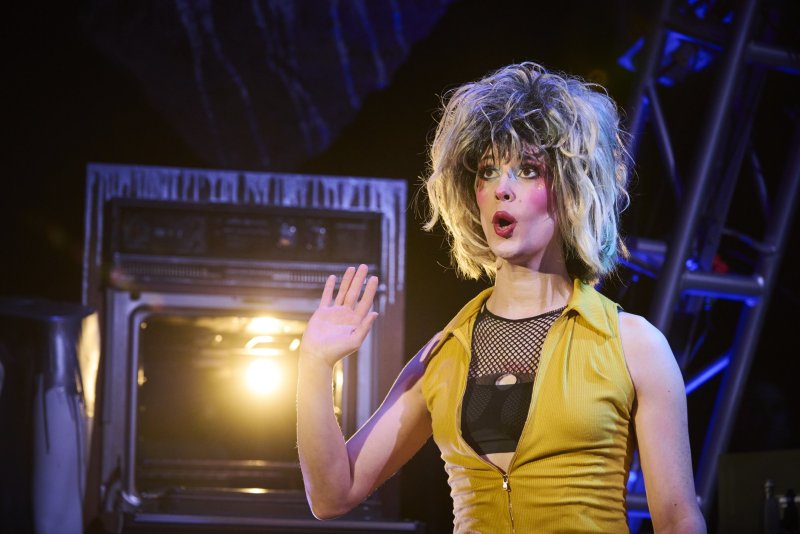 Performer in a yellow top, fishnet shirt, and blonde wig with dramatic makeup, mouth open, hand raised, on a dark stage.