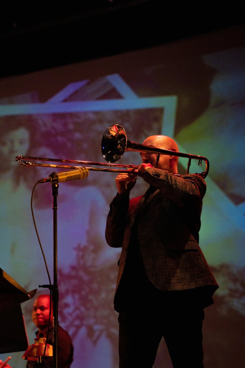 Bald man in a dark suit playing a trombone on stage, with a microphone in front and a blurry, colorful projection behind him.