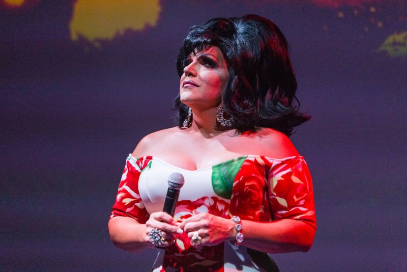 Drag performer in off-shoulder floral dress, holding microphone. Black wig, heavy makeup, and large earrings. Stage lighting with blurred background.