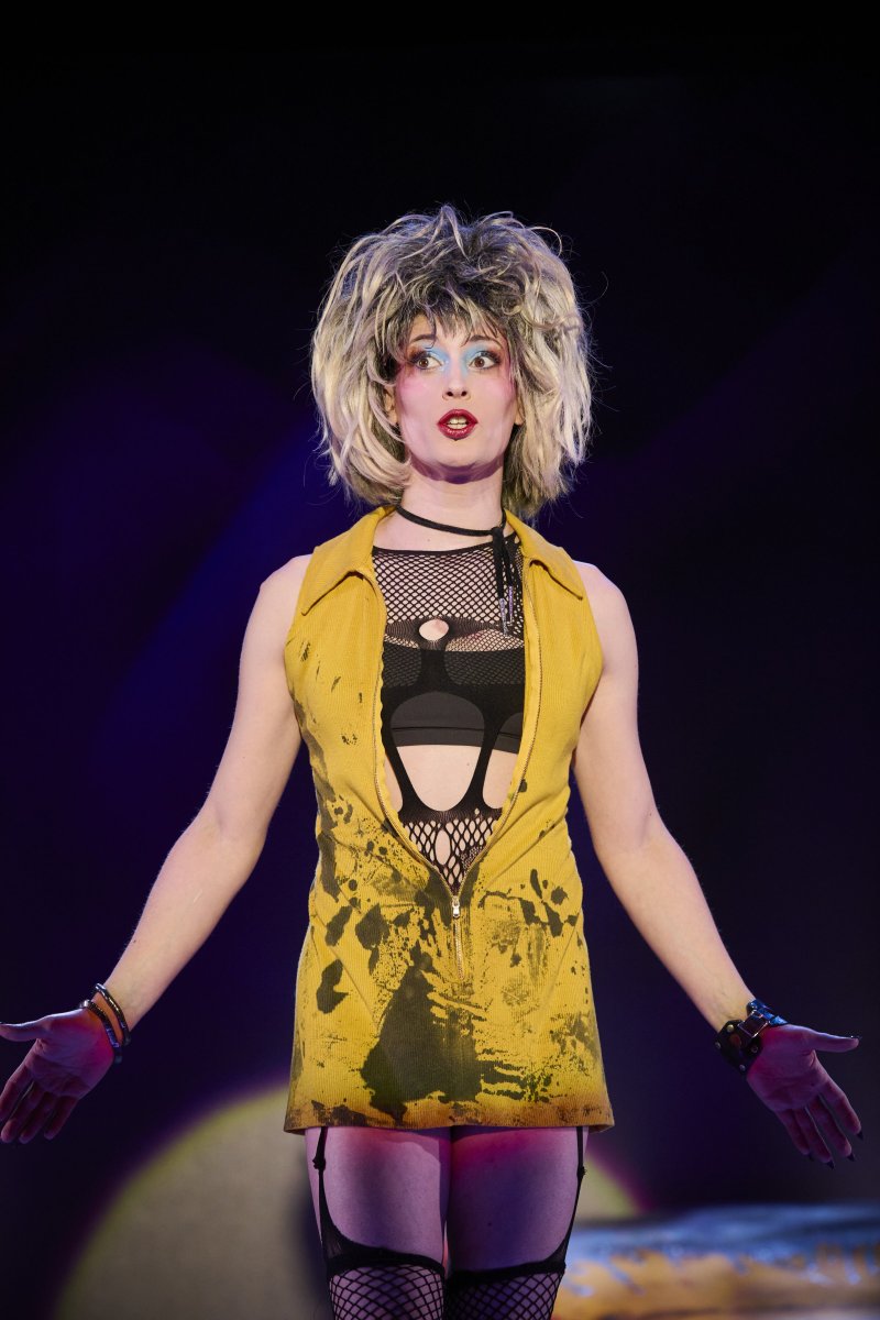 Drag performer with messy blonde wig, heavy makeup, yellow vest, fishnets, and black undergarments, arms outstretched on a dark stage.