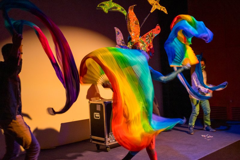 Drag performer Darrell Thorne in a vibrant rainbow costume with two assistants waving colorful fabrics on a dark stage.