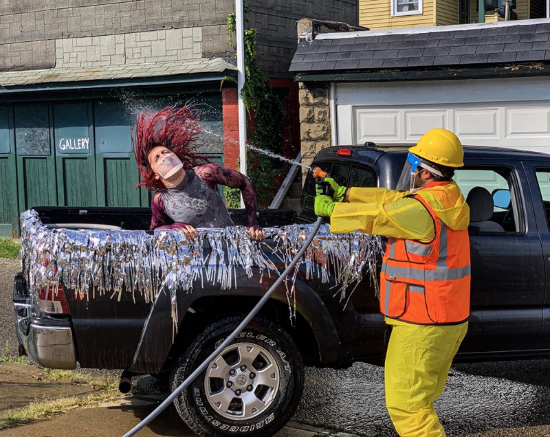 A person with red hair in a truck bed covered in tinsel is sprayed with water by a person in a yellow suit and hard hat.