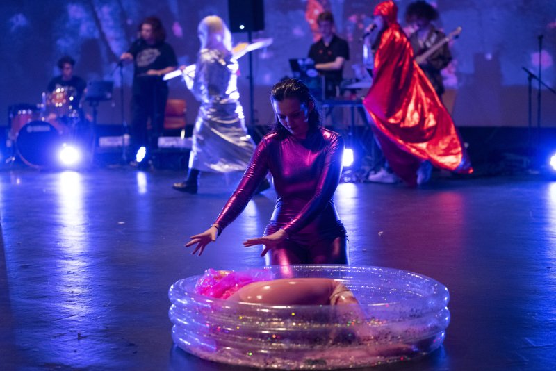 Performer in a shiny pink suit bending over a clear inflatable pool with a mannequin, band in background.