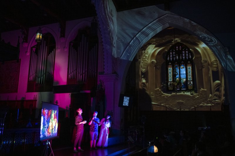Three figures bathed in pink and blue light stand before a large projection of a gothic window in a dark church interior.