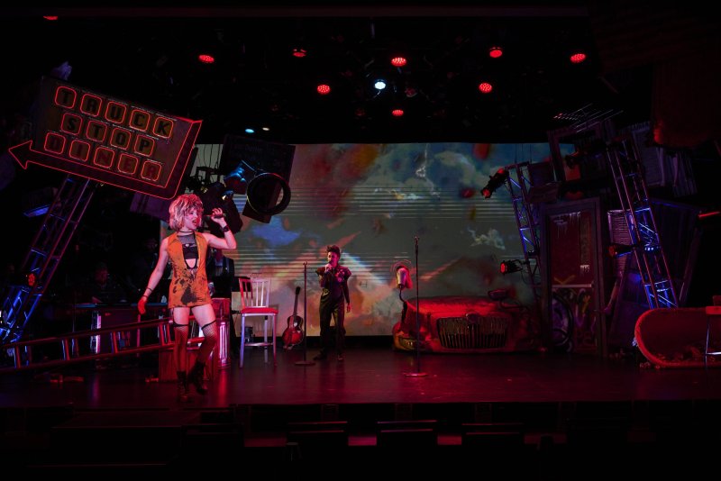 Two performers on a stage with a large screen displaying abstract art, surrounded by props and red stage lights.