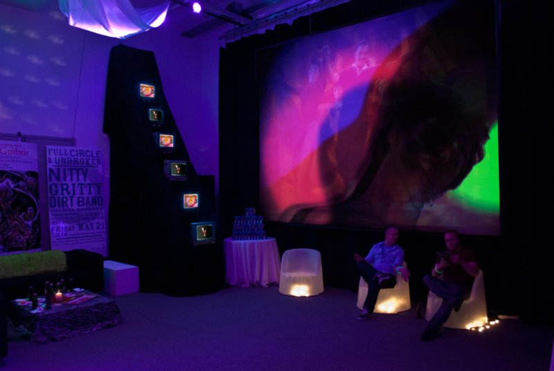 Art installation with two men seated on glowing chairs, facing a large screen displaying abstract colorful projections.