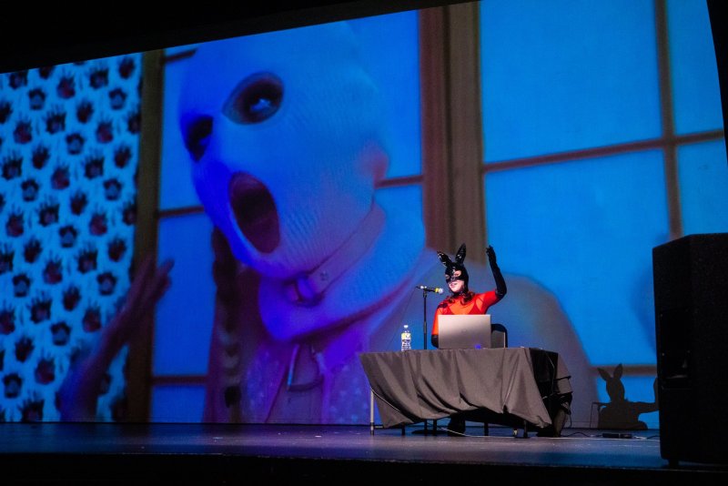 Performance art on stage: a woman in a red outfit and bunny mask stands at a table with a laptop, microphone and water bottle. Behind her, a large screen displays a close-up of a person in a white ski mask with an open mouth.