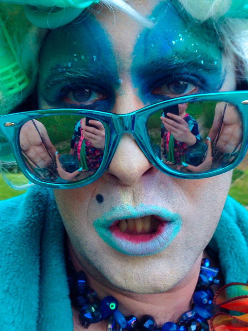 Close-up of a person in drag with blue and white makeup, turquoise sunglasses, and a blue necklace.