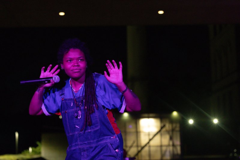 Performer in denim overalls, lit in purple, holds a microphone and raises hands. Dark background with blurred lights.