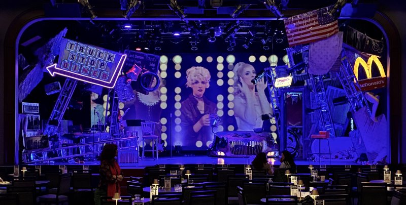 Stage set with neon "TRUCK STOP DINER" sign, American flag, McDonald's sign, and projected images of two performers.