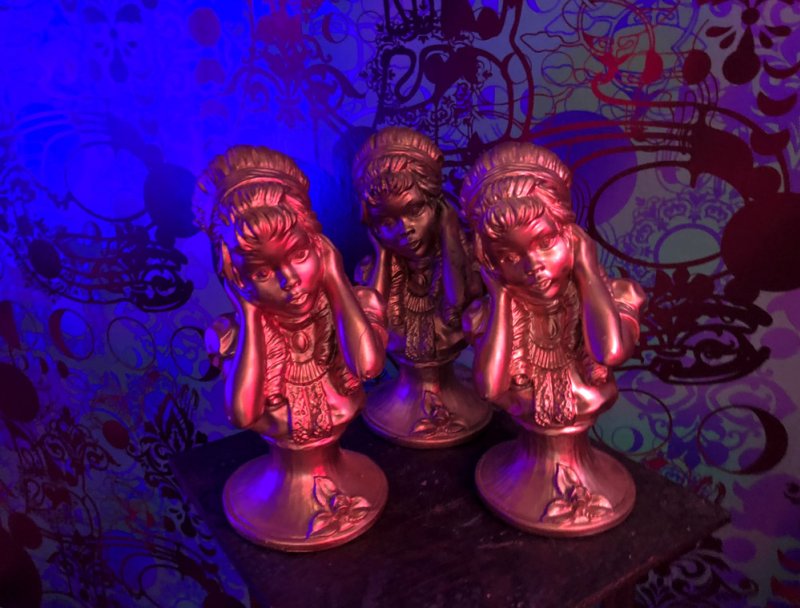 Three gold-painted busts of young girls with hands covering their ears, against a blue and purple patterned background.