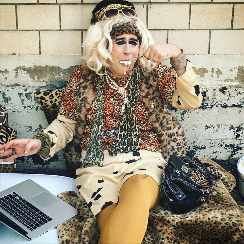 Drag queen in leopard print, blonde wig, and sunglasses, eating with a spoon, laptop nearby.