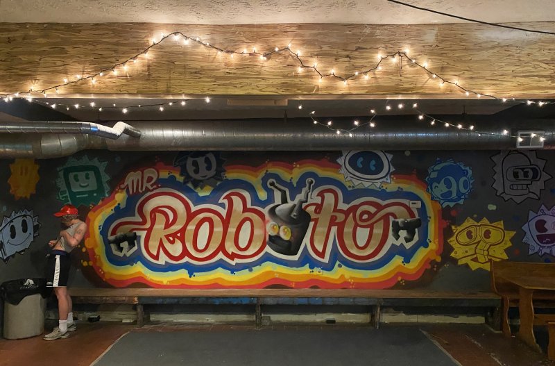 Graffiti art on a dark wall with "Mr. Roboto" in colorful bubble letters, surrounded by cartoon robots. A person stands left.