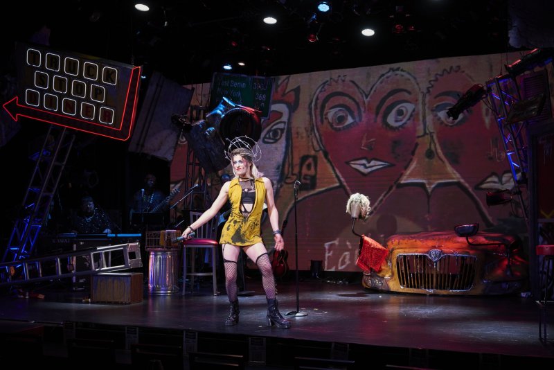 Drag queen in yellow dress, fishnets, and elaborate headpiece on stage with a vibrant, graffiti-style backdrop and neon signs.