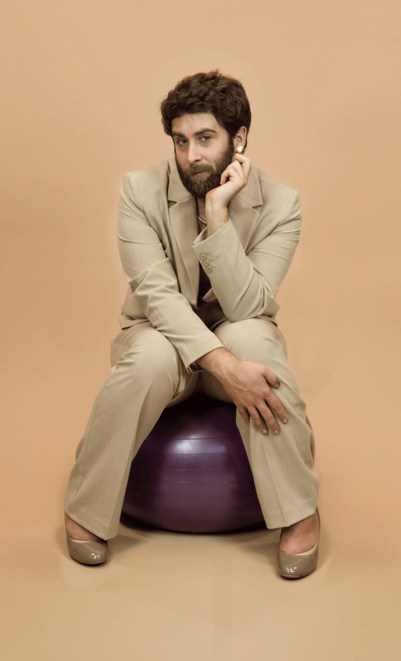 A bearded man in a beige suit and heels sits on a purple exercise ball, hand on chin, against a peach background.