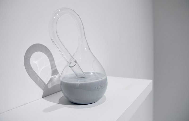 Clear glass vessel with a curved handle and spout, containing light gray liquid, casting a shadow on a white wall.