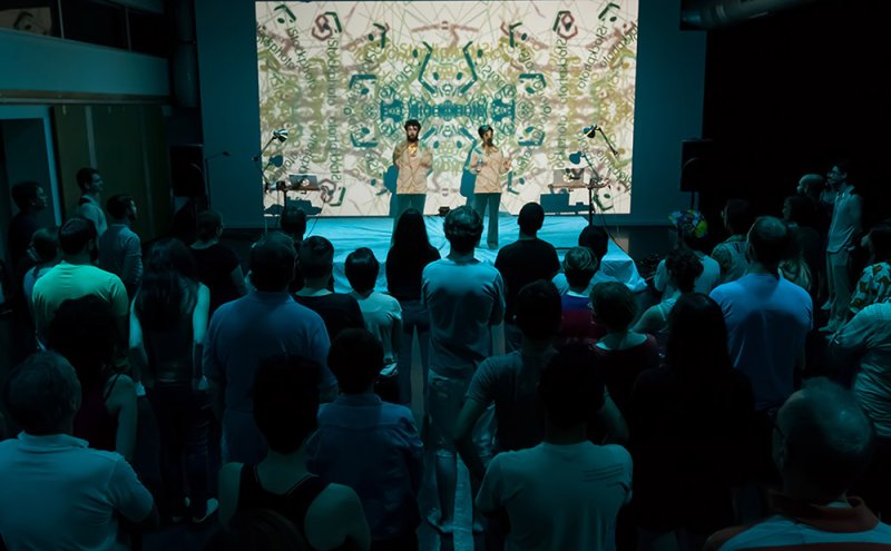 Performance art with two men on stage, projected onto a screen with abstract patterns, viewed by a dark audience.