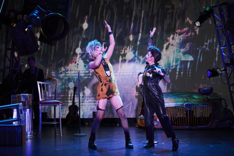 Two performers in drag on stage, one in a yellow dress and blonde wig, the other in black leather with dark hair, arms raised.