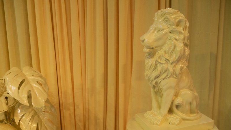 Monochromatic artwork features a glossy white lion sculpture, glossy white monstera leaves, and a gold curtain backdrop.