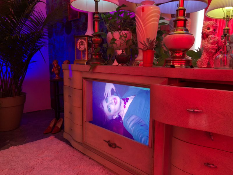 Installation view of a dresser with a screen displaying a person, lit in blue and red. Lamps, plants, and decorative objects sit on top. A rug and potted plant are nearby.