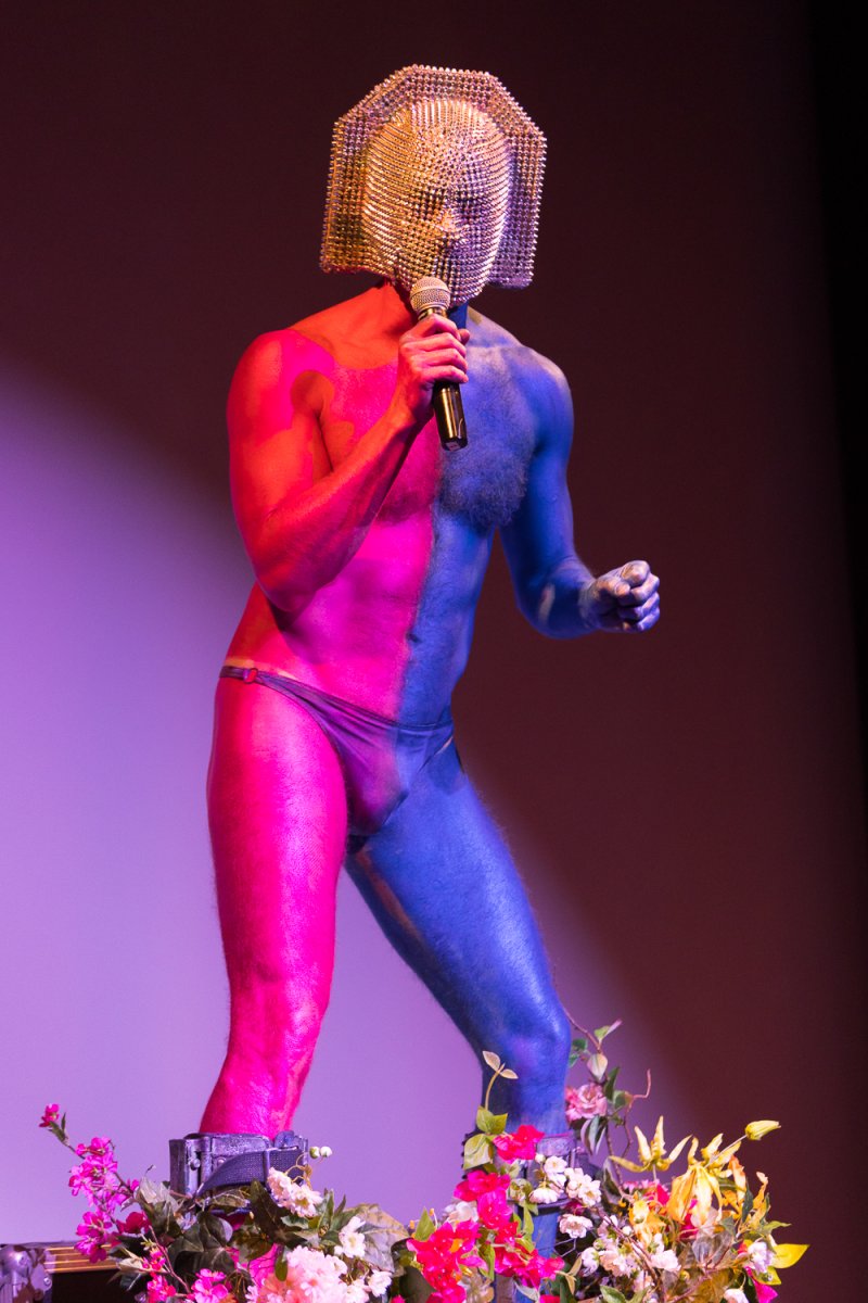 Performer Darrell Thorne, half-painted hot pink, half-painted blue, wearing a golden beaded mask and holding a microphone.