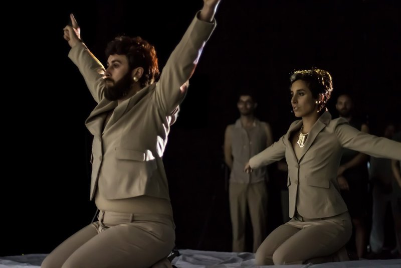 Two performers in beige suits, one bearded, kneel with arms raised on a white surface in a dark space.