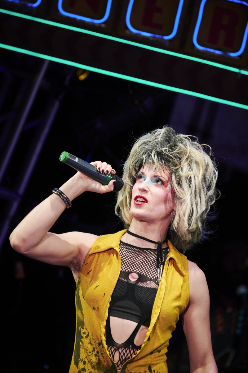 Performer with teased blonde hair, blue eyeshadow, and red lipstick, wearing a yellow vest and black mesh top, holding a microphone.