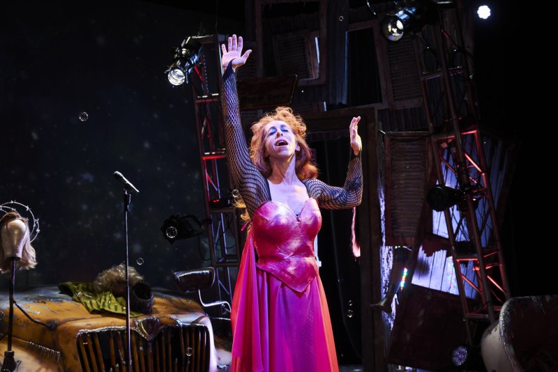 A performer in a shiny pink dress and fishnet sleeves sings with arms raised on a dark stage with a microphone and props.