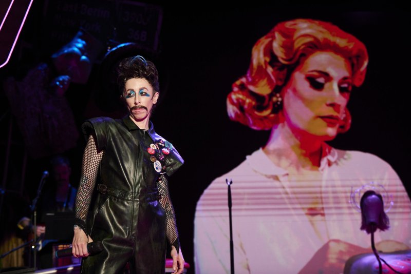 Performer in black leather and fishnets with painted mustache and blue eyeshadow, beside a projected image of a woman with red hair.