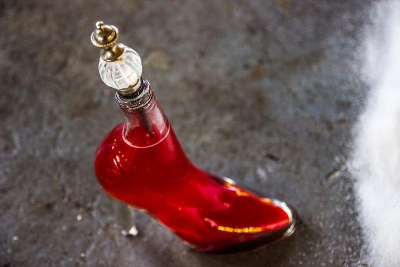 Glass high-heeled shoe bottle filled with red liquid, a clear and gold stopper, on a textured gray surface.