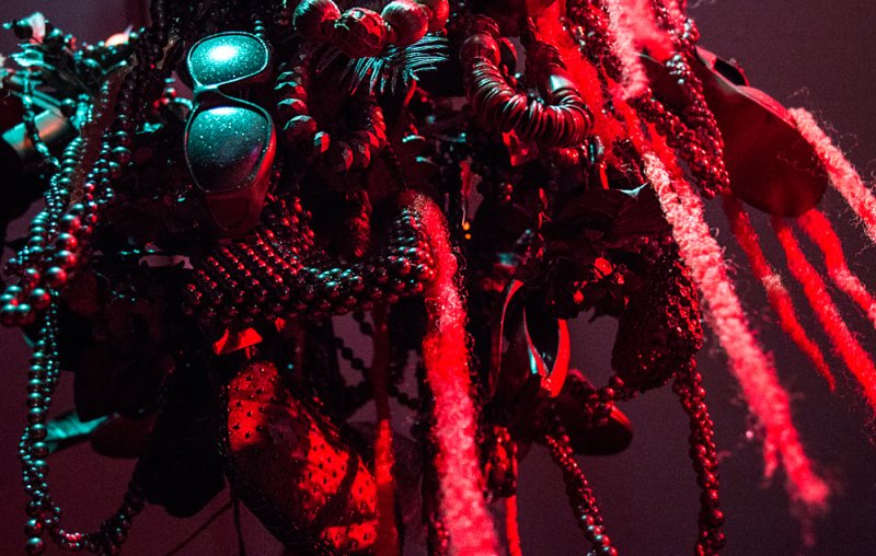 Close-up of a dark, intricate sculpture with beads, wires, and textured elements, illuminated by strong red and cyan lights.