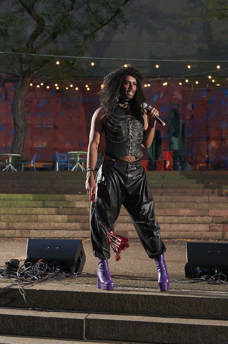 Performer on outdoor stage holds microphone, wearing leather vest with chains, baggy pants, purple platform boots, and holding red tassels. Speakers flank the stage. Background of trees and string lights.