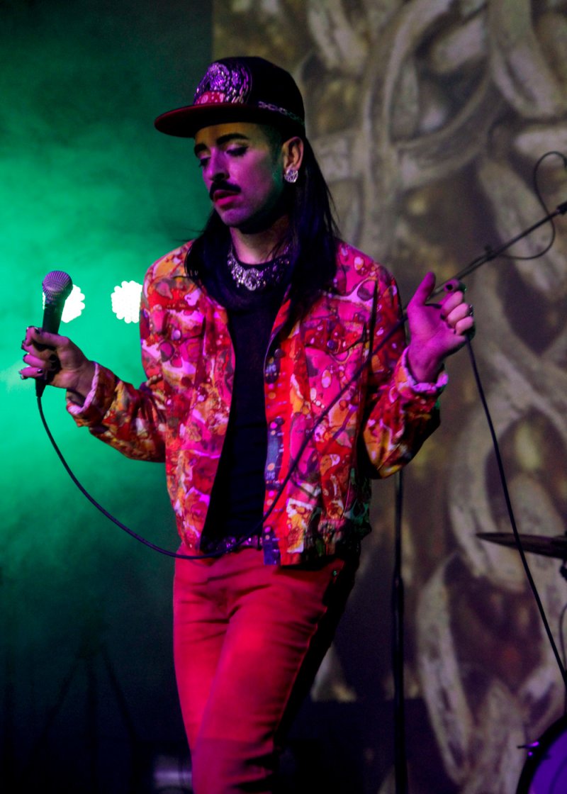 Performer in red pants and patterned jacket holds microphone in a green-lit space. They wear a black cap, necklace, and have a mustache. Background is blurred with abstract shapes.