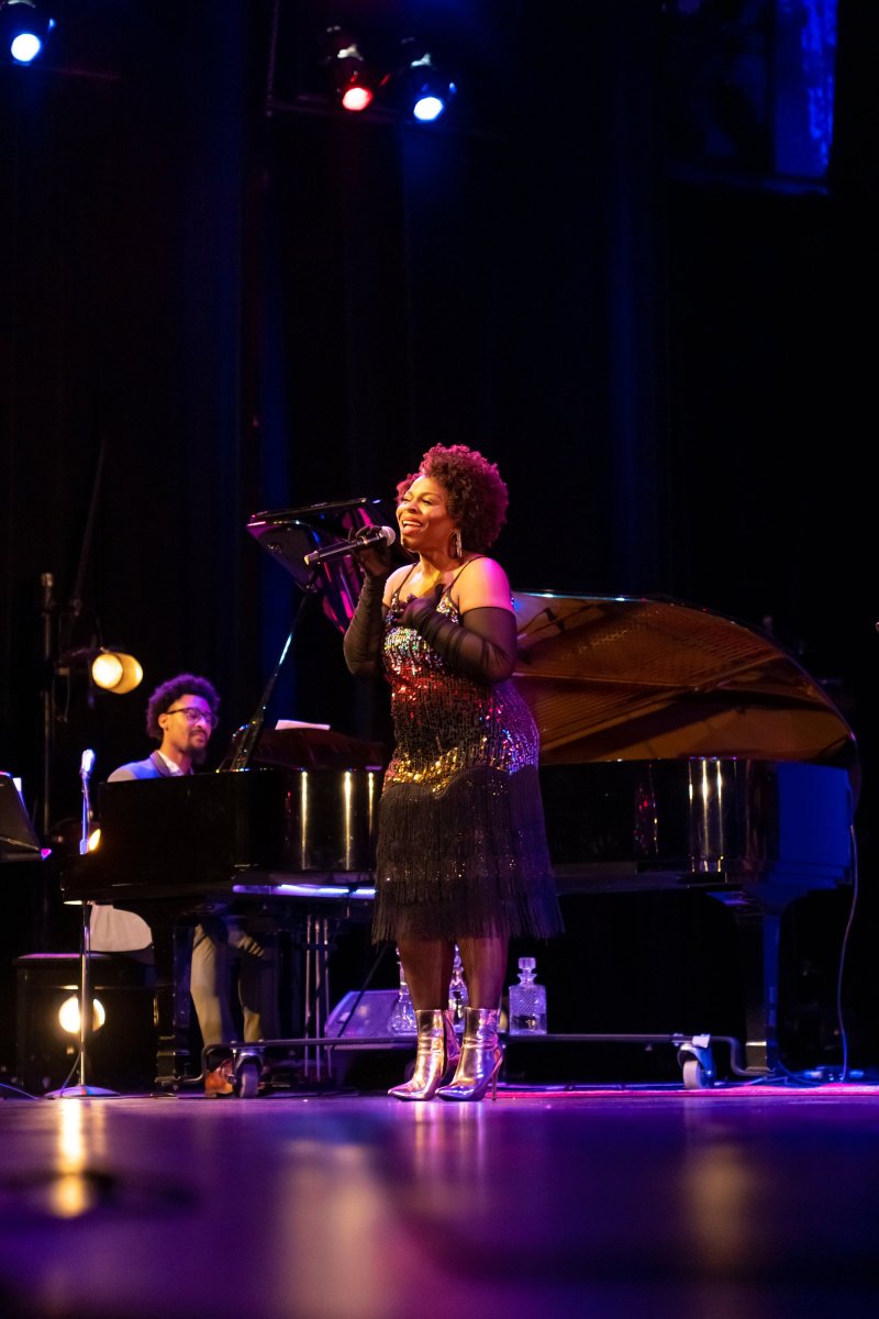 A Black woman sings on stage in a sequined dress and silver boots, a man plays piano behind her.