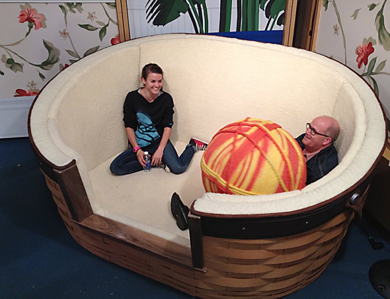 Two people in a giant cat bed with a huge yarn ball. A woman smiles, a man reclines.