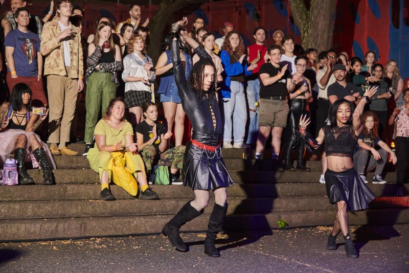Outdoor performance with two dancers in black leather outfits, one with dreadlocks, performing for a large crowd seated on stone steps. Colorful graffiti visible in the background.