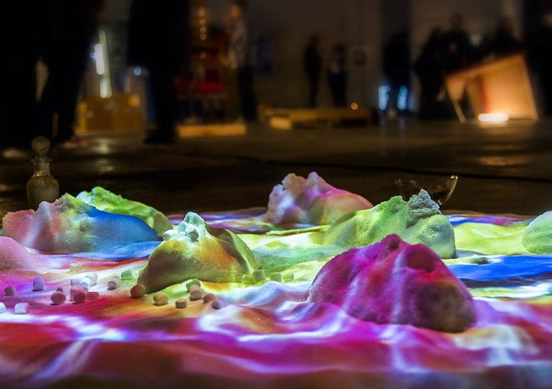 Installation view of sugar mounds lit with projected vibrant colors (pink, blue, green, yellow) on a dark floor. Small candies and glass vials are scattered around.