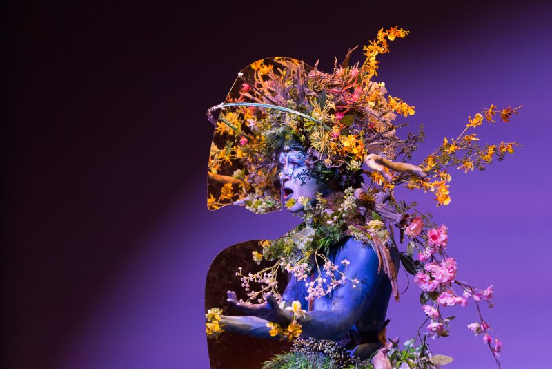 Drag artist Darrell Thorne in blue body paint, elaborate floral headpiece, and mirrored collar against a purple background.