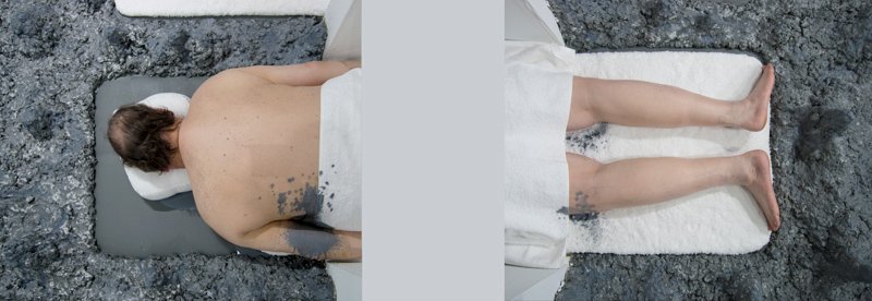 Two people lie face down on white mats, partially covered in dark gray material, on a textured dark gray surface.