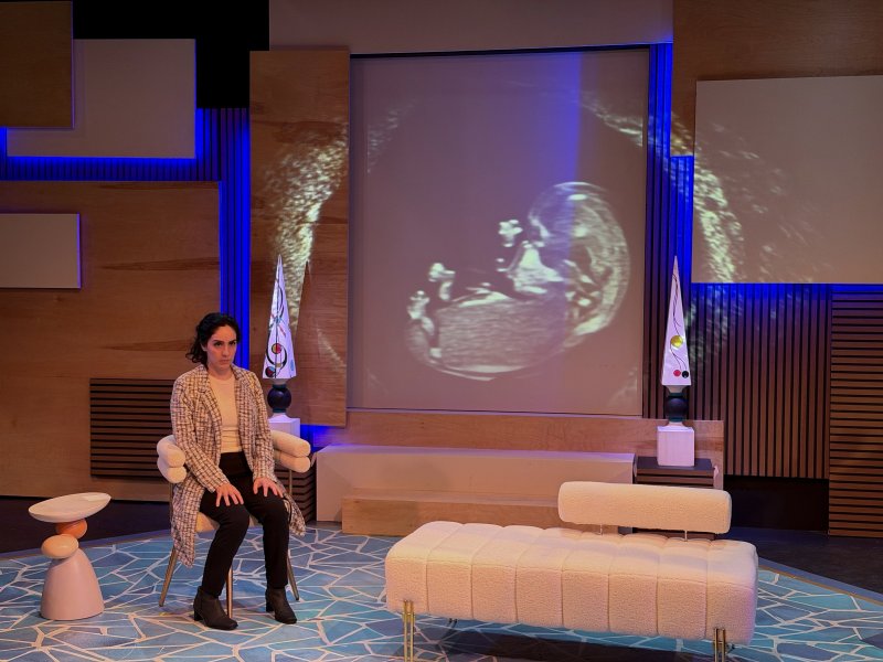 A woman sits on a chair on a stage with a fetal ultrasound projected on a large screen behind her.