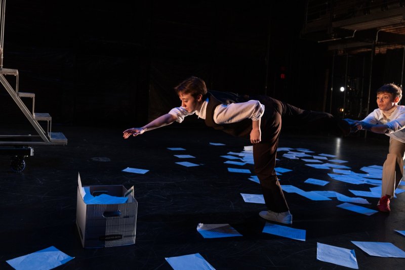 Two performers on a dark stage, one leaning forward with arm extended, the other pulling back; papers scattered on the floor.