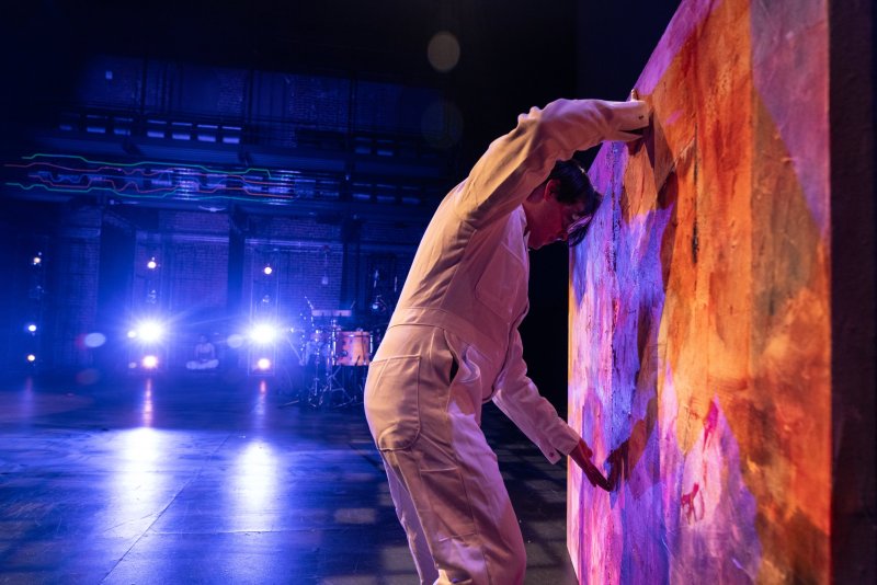Performer in white jumpsuit leans against a textured, abstract painting with warm tones, lit by cool blue stage lights.