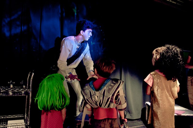 Stage performance with a man in a white and silver costume interacting with three children, one with bright green hair.